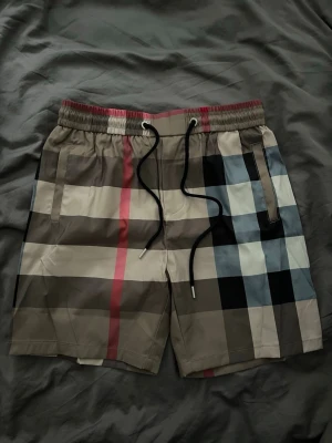 Burberry shorts - Snygga shorts med klassiskt rutigt mönster i beige, svart, blått och rött. Shortsen har elastisk midja med svart snörning och sidofickor samt en bakficka. Perfekta för sommaren och ger en trendig vibe.