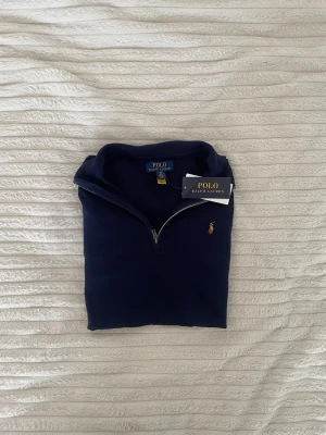 Mörkblå half-zip tröja Polo Ralph Lauren - Mörkblå långärmad tröja från Polo Ralph Lauren med klassisk half-zip och broderad logga på bröstet. Tillverkad i mjukt material och har ribbade muddar vid ärmslut och nederkant. Perfekt för en clean och stilren look.