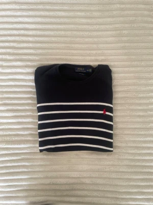Marinblå randig tröja Polo Ralph Lauren - Snygg marinblå stickad tröja från Polo Ralph Lauren med vita horisontella ränder och den klassiska röda loggan broderad på bröstet. Tröjan har rund halsringning och långa ärmar. Perfekt till jeans eller kjol för en stilren look.