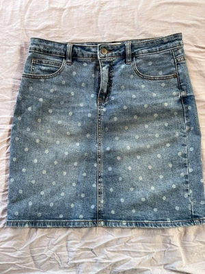 Blå jeanskjol med prickigt mönster - Säljer en blå jeanskjol med vita prickar och naturlig slitage look. Kjolen har bälteshällor, knapp och dragkedja framtill samt två bakfickor. Materialet är denim och kjolen har en rak passform som slutar ovanför knät.Aldrig använd!