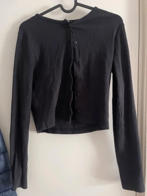 Svart ribbad cardigan med knappar - Trendig svart ribbad cardigan med långa ärmar och knäppning framtill. Kortare modell som passar perfekt till high waist jeans eller kjol. Tunn och mjuk i materialet, enkel att styla till olika outfits.