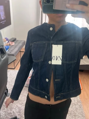 Mörkblå jeansjacka från Zara - Snygg mörkblå jeansjacka från Zara med raka linjer och silverfärgade knappar. Jackan har klassiska sömmar, rund halsringning och två bröstfickor. Perfekt för dig som gillar en stilren och trendig look.