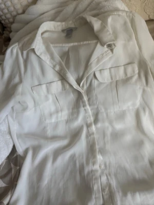 Vit skjorta från H&M med fickor - Klassisk vit skjorta från H&M med två bröstfickor och knappar framtill. Skjortan har trekvartsärm och är tillverkad i ett mjukt, lätt material som ger en avslappnad känsla. Perfekt att styla till jeans eller kjol.