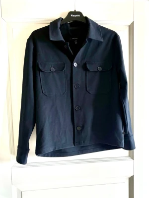 Mörkblå overshirt från Martinique  - Stilren mörkblå overshirt från Matinique med klassisk krage, fyra knappar framtill och två bröstfickor med lock och knapp. Tillverkad i ett mjukt bomullstyg som ger en avslappnad men snygg look. Perfekt att bära öppen eller stängd över en t-shirt. Använda ungefär 10 gånger. Nypris runt 1300 kr. Hör gärna av er vid frågor eller funderingar! 