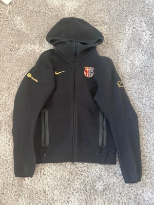 Nike Tech FC Barcelona - Svart hoodie från Nike med FC Barcelona-logga på bröstet och gul Nike Swoosh. Dragkedja framtill, två fickor med dragkedja och tryck på ärmarna, bland annat Spotify-logga. Perfekt för dig som älskar fotboll och vill ha en sportig look.