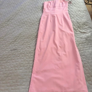 Ljusrosa långklänning med smyckade band - Supersnygg långklänning i ljusrosa med avtagbara axelband som har blommiga smyckesdetaljer. Klänningen är ärmlös och har en rak, elegant siluett. Materialet känns mjukt och lyxigt, perfekt för bal eller student.