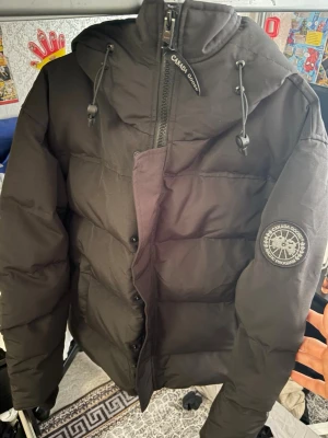 Svart dunjacka från Canada Goose - Snygg svart dunjacka från Canada Goose med huva och dragsko. Jackan har en tydlig logotyp på ärmen och grov dragkedja framtill med vindslå. Perfekt för kalla vinterdagar och riktigt varm tack vare dunfyllningen.