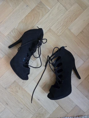 Svarta snörade stövletter med klack - Catwalk - Svarta stövletter med hög klack och öppen tå. Skorna har snörning framtill och dragkedja bak för enkel påtagning. Mockaimitation. Använd endast en gång inomhus.