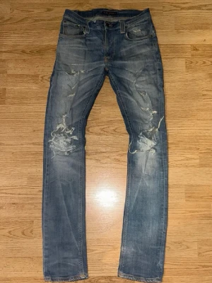 Nudie jeans ljusblåa - Säljer ett par blå jeans från Nudie Jeans med slitna detaljer och hål på benen. Jeansen har klassisk femficksdesign, normal passform och är tillverkade i bomull. Perfekta för en avslappnad och trendig look.