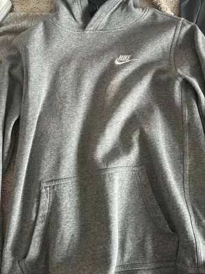 Grå hoodie från Nike - Snygg grå hoodie från Nike med klassisk logga på bröstet. Hoodien har huva och en stor magficka framtill. Tillverkad i mjukt sweatshirtmaterial som är skönt att ha på sig. Perfekt för en avslappnad och sportig stil.