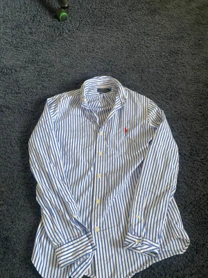Randig skjorta från Polo Ralph Lauren - Säljer en klassisk vit och blå randig skjorta från Polo Ralph Lauren i slim fit. Skjortan har långa ärmar, knappar framtill och den ikoniska röda loggan broderad på bröstet. Perfekt för en clean och stilren look. Hör av er vid frågor eller funderingar 