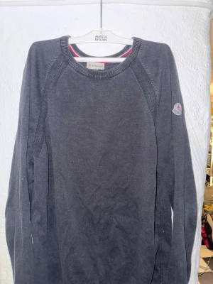 Mörkgrå stickad tröja från Moncler - Snygg mörkgrå stickad tröja från Moncler med rund halsringning och diskret Moncler-logga på ärmen. Tröjan har långa ärmar och ribbade muddar. Perfekt för dig som gillar stilrena och klassiska plagg.