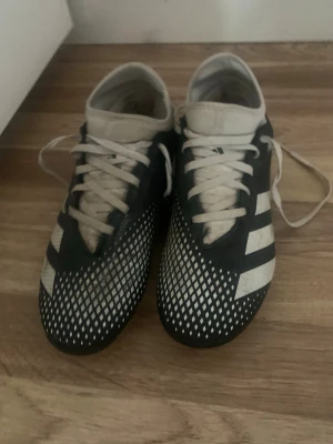 Adidas svartvita fotbollsskor - Säljer ett par klassiska Adidas fotbollsskor i svart och vitt med de ikoniska tre ränderna på sidan. Skorna har snörning och ett mönstrat material framtill för bättre bollkontroll. Perfekta för dig som vill ha stilrena och funktionella skor på planen.