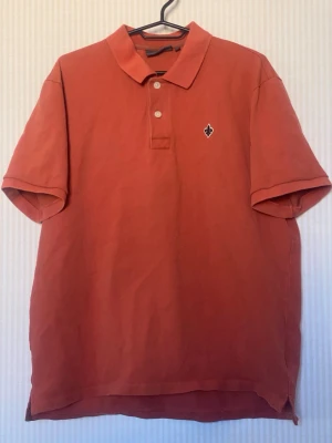 Rostfärgad pikétröja med krage - Snygg rostfärgad pikétröja med klassisk krage och två knappar framtill. Tröjan har korta ärmar och en liten broderad logga på bröstet. Perfekt till jeans eller shorts för en avslappnad stil.