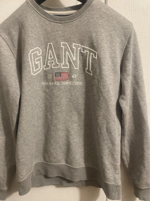 Koszula GANT / Grå sweatshirt från GANT - Klassisk grå sweatshirt från GANT med tryckt logga och amerikansk flagga på bröstet. Rund halsringning, ribbade muddar vid ärmslut och nederkant. Perfekt för en avslappnad och stilren look. Tröjan är i barnstorleken 170 som motsvarar ungefär storlek S för män. 