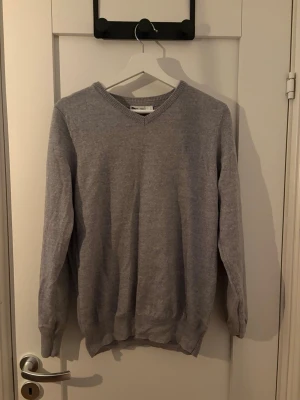 Ljusgrå stickad tröja - En stilren ljusgrå stickad tröja med V-ringning och långa ärmar. Tröjan har ribbade muddar vid ärmslut och nederkant, vilket ger en snygg passform. Perfekt att matcha med jeans eller kjol för en enkel och clean look.