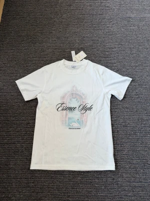 Vit Essence Style t-shirt - Nypris 790kr. Vit t-shirt från Fountain of Savory med trycket 'Essence Style' och ett motiv av en svan och ett dekorativt valv i pastellfärger. Klassisk passform och rund hals. Perfekt för dig som gillar stilrena men ändå unika plagg.