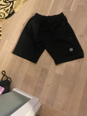 Svarta shorts från Stone Island - Säljer ett par svarta shorts från Stone Island med elastisk midja och snörning. Shortsen har en avslappnad passform och klassisk Stone Island-logga på benet. Perfekta för chill dagar och enkel streetwear-stil.