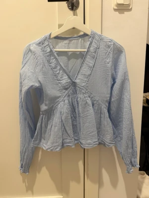 Ljusblå omlottblus med volang - Supersöt ljusblå blus med omlott-look och volang nedtill. Blusen har v-ringning och långa ärmar med lätt rynk vid ärmslut. Tillverkad i tunn, luftig bomull som känns skön mot huden. Perfekt för dig som gillar en feminin och avslappnad stil.