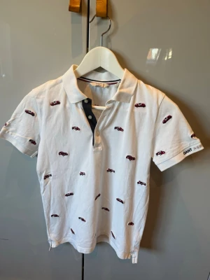 Vit pikétröja med bilbrodyr från GANT - Vit pikétröja från GANT med små broderade bilar i rött och svart över hela tröjan. Klassisk krage, korta ärmar och knappslå framtill. GANT-logga broderad på ärmen. Perfekt för en avslappnad men snygg stil.