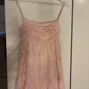 Ljusrosa kort klänning med rosett - Söt ljusrosa kort klänning med tunna axelband och volangdetaljer. Klänningen har en dekorativ rosett i ryggen och ett subtilt blommigt mönster i tyget. Perfekt för varma dagar och somriga tillfällen.