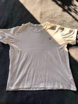 Vit t-shirt från GANT med logga - Klassisk vit t-shirt från GANT med liten broderad logga och sköld på bröstet. T-shirten har rund halsringning och korta ärmar. Tillverkad i mjuk bomull som känns skön mot huden. Perfekt basplagg för alla tillfällen.