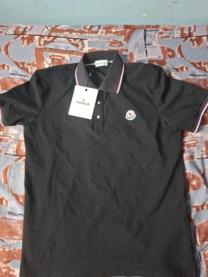 Svart Moncler pikétröja med krage - Klassisk svart pikétröja från Moncler med vit och röd detalj på kragen och ärmsluten. Tröjan har korta ärmar, knappslå framtill och Moncler-logga på bröstet. Perfekt för en stilren och sportig look.