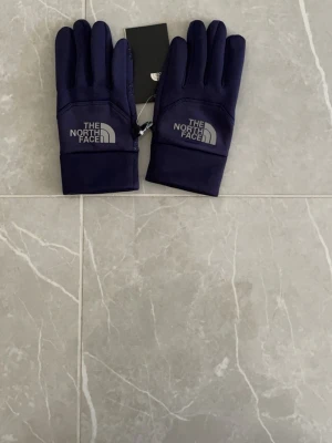 Navy blue handskar från The North Face - Snygga mörkblå handskar från The North Face med logga på ovansidan. Handskarna har greppvänligt mönster i handflatan och fingertoppar anpassade för touchskärm. Perfekta för kalla dagar och outdoor-aktiviteter.