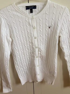 Vit kabelstickad kofta från Gant - Kabelstickad vit kofta från Gant i stretchig bomull. Klassisk rund halsringning, knäppning framtill med vita knappar och diskret Gant-logga broderad på bröstet. Perfekt för en clean och stilren look.