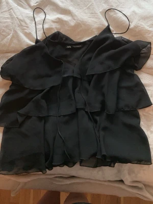 Svart volangblus från Zara - Säljer en svart blus från Zara med tunna axelband och flera lager volanger framtill. Knyte som man kan knyta vid bysten. Sparsamt använd. 