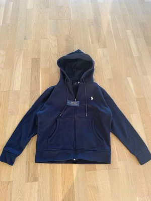 Mörkblå hoodie från Polo Ralph Lauren - Snygg mörkblå hoodie från Polo Ralph Lauren med dragkedja framtill och klassisk vit logga på bröstet. Hoodien har två stora fickor fram och justerbar huva med snören. Tillverkad i mjukt bomullsmaterial som känns skönt mot huden.