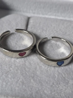 Justerbara silvriga ringar med hjärtan bff  - Två stilrena silvriga ringar i metall med små emaljerade hjärtan framtill, en i blått och en i rosa. Ringarna är justerbara och har en enkel, modern design som passar perfekt för att matcha med andra smycken eller bära var för sig. De lyser lite lätt i mörker efter att ha fått solljus på sig.