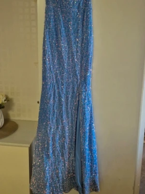 Blå glittrig långklänning - Säljer en blå långklänning med glittriga paljetter över hela tyget. Klänningen har en elegant slits framtill och är perfekt för speciella tillfällen. Materialet känns festligt och skimrande, och modellen är figurnära med en klassisk siluett.
