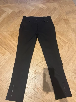 Svarta kostymbyxor med knappar - Snygga svarta kostymbyxor med smal passform och dekorativa knappar vid bensluten. Byxorna har en klassisk midja, bälteshällor och fickor fram. Perfekta för en stilren look. Materialet ser ut att vara en mjuk och stretchig mix, vilket ger skön komfort.