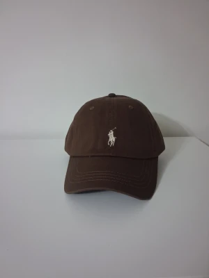 Brun keps från Polo Ralph Lauren - Snygg brun keps från Polo Ralph Lauren med klassisk böjd skärm och justerbart spänne bak. Framtill finns den ikoniska vita polospelaren broderad och baktill står det 'POLO' broderat i vitt. Tillverkad i bomull för en bekväm känsla.