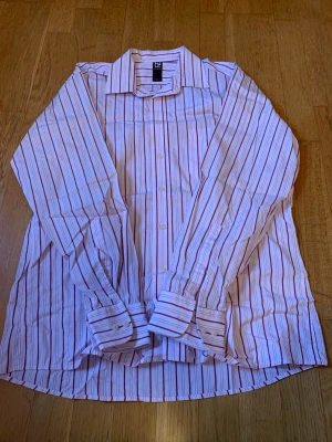 Randig skjorta från LE LAB Black Label - Säljer en vit skjorta med tunna röda och lila ränder från LE LAB Black Label. Skjortan har klassisk krage, knäppning framtill och långa ärmar med manschetter. Perfekt för dig som gillar stilrena och tidlösa plagg.