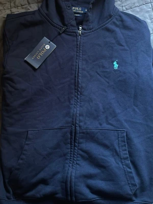 Mörkblå hoodie från Polo Ralph Lauren - Snygg mörkblå hoodie från Polo Ralph Lauren med dragkedja framtill och klassisk huva. Märkets ikoniska logga i ljusblått broderad på bröstet. Hoodien har känguruficka och är tillverkad i mjukt bomullsmaterial. Perfekt för en avslappnad och stilren look.