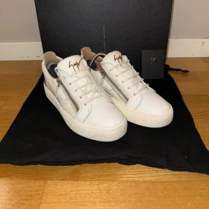 Vita sneakers från Giuseppe Zanotti - Snygga vita sneakers från Giuseppe Zanotti med silverfärgade dragkedjor på sidorna och logga i metall på plösen. Skorna har en låg siluett, vita snören och är tillverkade i slätt läder. Perfekta för dig som gillar stilrena och exklusiva detaljer.