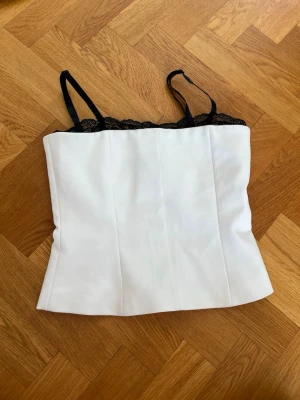 Vit korsettopp med spetsdetalj - Snygg vit korsettopp med svarta justerbara axelband och svart spets längs överkanten. Toppen har en figurnära passform och är perfekt för att lyfta vilken outfit som helst. Materialet känns slätt och har en lätt struktur.