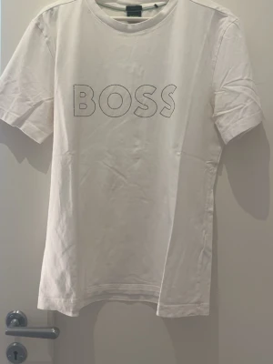 Vit BOSS t-shirt med tryck - Snygg vit t-shirt från BOSS med klassiskt rund halsringning och korta ärmar. Framsidan pryds av ett stort BOSS-tryck i svart. Tillverkad i mjuk bomull för skön känsla och enkel stil.