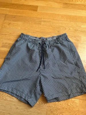 Mönstrade badbyxor från H&M - Säljer ett par snygga badbyxor från H&M i storlek M. De har ett blåvitt grafiskt mönster, elastisk midja med dragsko och meshfoder på insidan. Perfekta för stranden eller poolen och riktigt bekväma att bära.