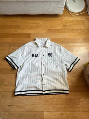 Vit randig skjorta från Bershka - Säljer en vit skjorta från Bershka med svarta tunna ränder och tryck 'MIA 98' på bröstet. Skjortan har korta ärmar, knappar framtill och sportig basebollstil. Perfekt för dig som gillar streetwear och vill sticka ut.