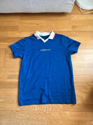 Blå pikétröja från Iets Frans - Säljer en blå pikétröja från Iets Frans med vit krage och röd detalj på kragen. Tröjan har korta ärmar och broderad logga på bröstet. Materialet är mjukt och lätt, perfekt för en avslappnad stil.