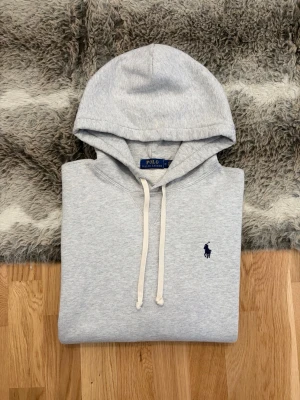 Grå Hoodie Från Polo Ralph Lauren - Storlek M - Hej! 👋 Säljer en klassisk grå Polo Ralph Lauren hoodie i storlek M. Mörkblå pony-logga broderad på bröstet 🐎 Tröjan är i gott skick överlag med mjuk fleece-insida, men observera att det finns en mindre blekning/fläck över magfickan (se bild) ✨ Sitter kanon på (Modell: 177cm) 👕 Modellen saknar QR-kod men alla invändiga tvättlappar sitter kvar för att garantera äktheten ✅ Perfekt för dig som vill ha ett snyggt märkesplagg till ett lite bättre pris! Fraktar snabbt 🚛📦💬💯 
