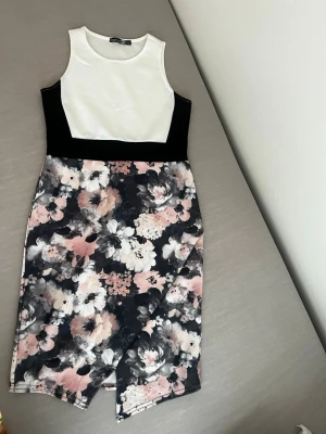 Blommig klänning från Boohoo - Snygg ärmlös klänning från Boohoo med vit överdel och svart midja. Kjoldelen har ett blommigt mönster i svart, vit och rosa med asymmetrisk nederkant. Tillverkad i mjuk polyester med normal passform. Perfekt för dig som vill sticka ut lite extra.