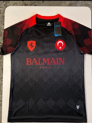 L Turkisk svart och röd fotbollströja  - Med pris lapp Bara köpte det va stor på mig har M trodde den skulle passa den är L Snygg svart och grå  med röda detaljer och balmain tryck. Tröjan har korta ärmar, grafiskt mönster, och loggor på bröstet som turkisk flagga och Ferrari På ryggen står det ASLAN, vilket betyder lejon på turkiska mit eget köpt design är hella tröjan nummer 7 och FLAWLESS i rött. Perfekt för träning eller match.