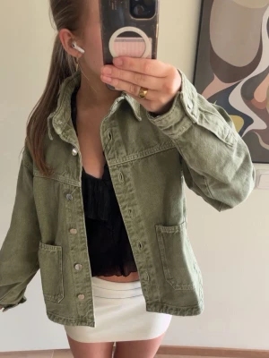 Olivgrön oversized jeansjacka - Snygg olivgrön jeansjacka i oversized modell med klassisk krage och tryckknappar framtill. Två stora fickor framtill och raka ärmar ger en avslappnad look. Perfekt att slänga över en topp för en trendig stil.
