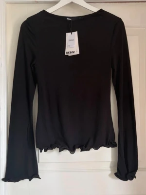Svart långärmad topp från Bik Bok - Svart långärmad topp från Bik Bok med volangkant vid ärmslut och nederkant. Tunn och mjuk i materialet, perfekt för att styla med jeans eller kjol. Enkel och stilren design med rund halsringning.