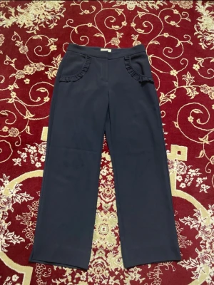 Mörkblå Lindex Kostymbyxor - Mörkblå byxor med raka ben och hög midja. Framtill finns två fickor med volangdetaljer som ger en unik touch. Använda ett par gånger men inga skavanker, bara en liten fläck på knät som ses på sista bilden. 2 för 150 kr på alla dambyxor!