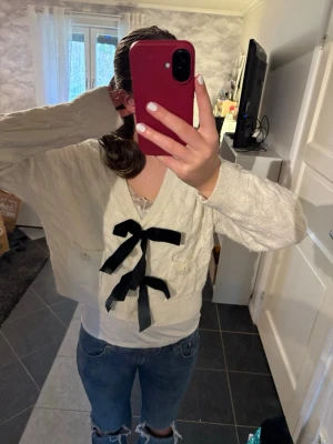 Vit stickad kofta med svarta rosetter - Söt vit stickad kofta med två stora svarta rosetter framtill. Koftan har v-ringning, kabelstickat mönster och ribbade muddar vid ärmslut och nederkant. Perfekt för dig som gillar en gullig och trendig stil.